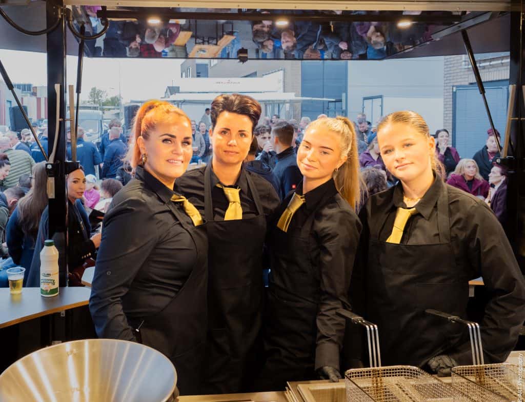 Gezellige medewerkers in zwart uniform met goudgele strik op een foodtruck tijdens evenement.