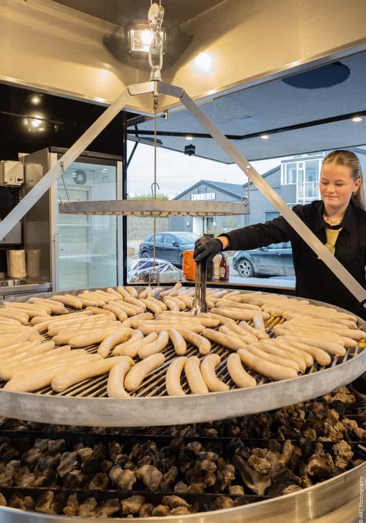 Gegrilde witte worst op een grote roterende grill, verzorgd door een medewerker.